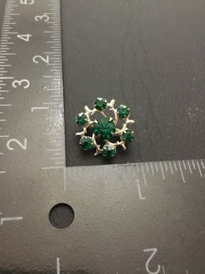 Sale Photo Thumbnail #144: Small vintage silver-tone wreath brooch featuring a center green faceted rhinestone surrounded by six smaller green rhinestones set in stylized branch-like prongs. Features a standard pin back with safety clasp closure. Measures approximately 0.75 inches 