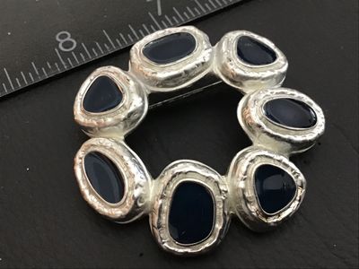 Sale Photo Thumbnail #87: A vintage circular wreath-style brooch featuring textured silver-tone metal set with dark blue/navy enamel or stone cabochons. The back features a crosshatched pattern with a copyright mark and a functional pin back clasp. Measures approximately 1.5 inche