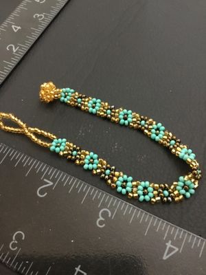 Sale Photo Thumbnail #124: Handmade woven seed bead bracelet featuring a turquoise-blue and gold-tone floral daisy chain design. Includes a beaded loop and beaded ball toggle closure. Measures approximately 7 inches long. In good condition with no missing beads visible.