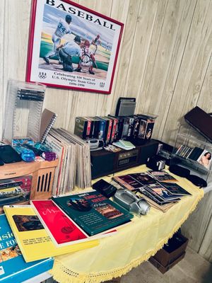 Collection of vintage media and home electronics displayed on a table, featuring a framed Olympic baseball poster, vinyl records, VHS...