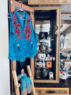 Vintage Western-style denim vest featuring red chili pepper embroidery and a fringed hem, hanging beside a display cabinet with...