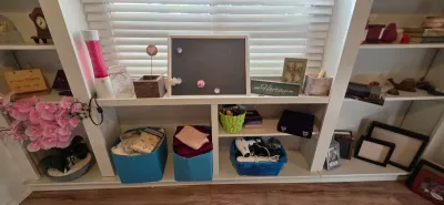Sale Photo Thumbnail #28: White built-in shelving unit holding assorted home decor, picture frames, magnetic board, artificial pink flowers, storage bins, and personal care appliances.
