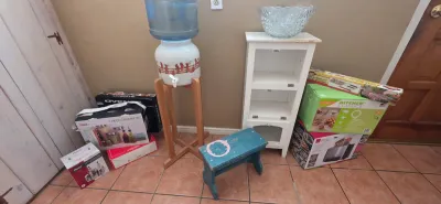Sale Photo Thumbnail #26: Collection of household goods including a ceramic water crock on a wooden stand, small painted blue footstool, white corner shelf, and various boxed kitchen appliances.