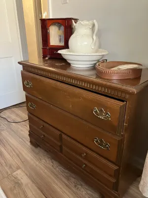 Sale Photo Thumbnail #43: Traditional wooden chest of drawers with bail handles, topped with a white ceramic pitcher and basin set, a wooden jewelry box, and a leather belt.
