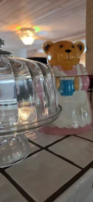 Sale Photo Thumbnail #4: Clear glass domed cake stand, positioned next to a vintage ceramic teddy bear cookie jar on a tiled kitchen counter.