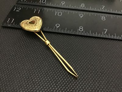 Sale Photo Thumbnail #78: Gold tone bodkin / ribbon threader featuring a hinged, opening heart-shaped locket top. The locket features floral and scroll engraved detailing on the front face. Measures approximately 3 inches in length. In good vintage condition with minor surface wea