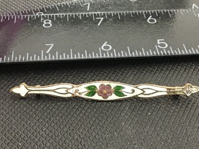 Sale Photo Thumbnail #202: Antique bar pin featuring a central hand-painted style enamel purple flower with green leaves framed by white enamel scrollwork on a brass or gold-tone metal base. Shows age-appropriate wear and patina, with light wear to the enamel finish. Measures appro
