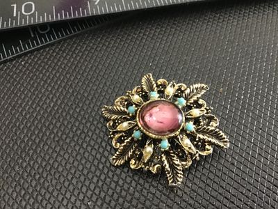 Sale Photo Thumbnail #211: Vintage gold-tone ornate filigree brooch featuring a central oval pink cabochon accented by faux pearls and turquoise-color beads. Features a safety pin clasp closure on the back. Shows light surface wear and finish aged patina consistent with age. Measur