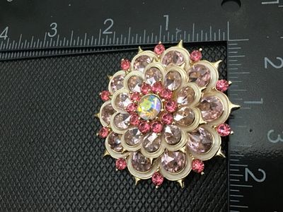 Sale Photo Thumbnail #31: Vintage gold-tone layered flower brooch featuring pale pink pear-cut rhinestones, dark pink round accent rhinestones, white enamel details, and a central iridescent aurora borealis rhinestone. Measures approximately 2 inches in diameter. Pin back with C-c