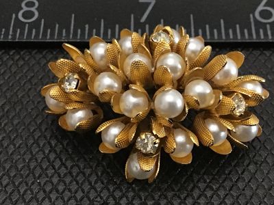 Sale Photo Thumbnail #95: A vintage gold-tone floral cluster brooch featuring faux pearls encased in textured, petal-like cups along with clear rhinestone accents. The pin measures approximately 1.5 inches in length. Features a standard pin back with a safety clasp mechanism. Exce
