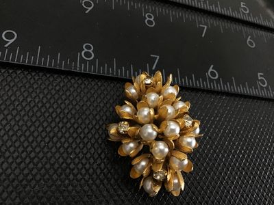 Sale Photo Thumbnail #94: A vintage gold-tone floral cluster brooch featuring faux pearls encased in textured, petal-like cups along with clear rhinestone accents. The pin measures approximately 1.5 inches in length. Features a standard pin back with a safety clasp mechanism. Exce