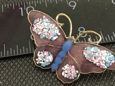 Sale Photo Thumbnail #182: A gold-tone butterfly brooch featuring purple translucent enamel wings accented with colorful pink, light blue, and white specks, with a blue body. Measures approximately 1.5 inches wide. Features a standard pin back with safety clasp in good working orde