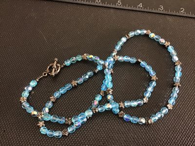 Sale Photo Thumbnail #232: A beaded necklace featuring light blue iridescent/aurora borealis faceted crystal beads, interspersed with silver-tone floral spacer beads and finished with a toggle clasp closure. Measures approximately 22 to 24 inches in total length. Overall good pre-o