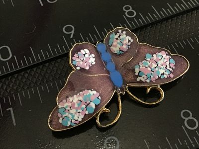 Sale Photo Thumbnail #184: A gold-tone butterfly brooch featuring purple translucent enamel wings accented with colorful pink, light blue, and white specks, with a blue body. Measures approximately 1.5 inches wide. Features a standard pin back with safety clasp in good working orde