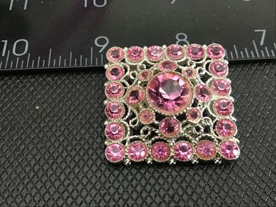 Sale Photo Thumbnail #175: A square-shaped vintage costume brooch featuring a central large round pink rhinestone surrounded by smaller, vibrant pink rhinestones set in silver-tone ornate filigree metalwork. Features a standard pin back with a safety clasp. Measures approximately 1