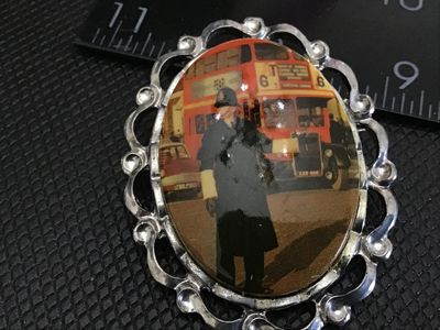 Sale Photo Thumbnail #174: An oval vintage brooch featuring a printed image of a London police officer (Bobby) in front of a red double-decker bus. Set in a silver-tone filigree metal frame with a pin-back closure. Shows light wear consistent with age.