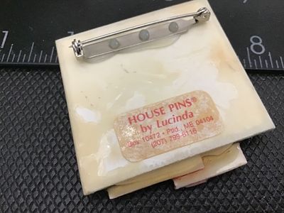 Sale Photo Thumbnail #72: A vintage artisan brooch labeled 'House Pins by Lucinda', Portland, ME. Features a colorful red house scene against a black background with gold glitter accents. Crafted with resin/plastic materials and a rollover safety clasp on the reverse. Measures app