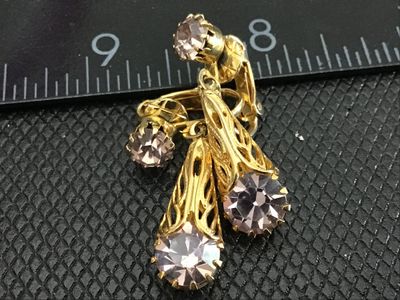Sale Photo Thumbnail #155: Pair of vintage gold-tone clip-on earrings featuring filigree cone drops set with light purple or amethyst-colored round rhinestones. Each earring has a smaller rhinestone set at the clip base with a dangling filigree component below. Measure approximatel