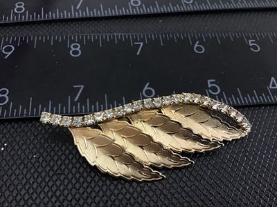 Sale Photo Thumbnail #145: A vintage gold-tone brooch featuring a cascading layered leaf motif with textured detail, accenting a curved line of clear set rhinestones along the top edge. The piece measures approximately 2.5 inches in length. Features a classic pin-back closure with 