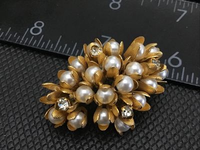 Sale Photo Thumbnail #91: A vintage gold-tone floral cluster brooch featuring faux pearls encased in textured, petal-like cups along with clear rhinestone accents. The pin measures approximately 1.5 inches in length. Features a standard pin back with a safety clasp mechanism. Exce