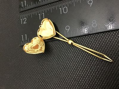 Sale Photo Thumbnail #76: Gold tone bodkin / ribbon threader featuring a hinged, opening heart-shaped locket top. The locket features floral and scroll engraved detailing on the front face. Measures approximately 3 inches in length. In good vintage condition with minor surface wea