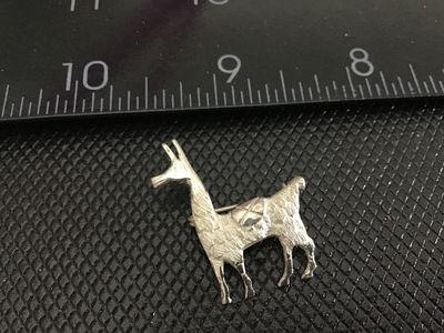Sale Photo Thumbnail #217: A silver-tone metal pin crafted in the shape of a llama or alpaca featuring textured fur detailing and a saddle back. Pin back fastener on the reverse. Measures approximately 1.25 inches long. Shows light patina and wear consistent with age.