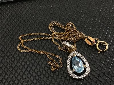 Sale Photo Thumbnail #179: Gold-tone fine link chain necklace featuring a teardrop/pear-cut light blue stone pendant surrounded by a textured silver-tone halo bezel. The pendant shows light wear on the back metal setting. Includes a spring ring clasp. Pendant measures approximately