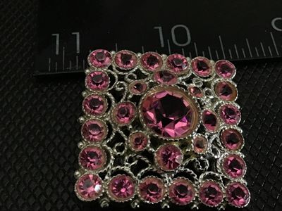 Sale Photo Thumbnail #178: A square-shaped vintage costume brooch featuring a central large round pink rhinestone surrounded by smaller, vibrant pink rhinestones set in silver-tone ornate filigree metalwork. Features a standard pin back with a safety clasp. Measures approximately 1