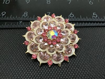 Sale Photo Thumbnail #30: Vintage gold-tone layered flower brooch featuring pale pink pear-cut rhinestones, dark pink round accent rhinestones, white enamel details, and a central iridescent aurora borealis rhinestone. Measures approximately 2 inches in diameter. Pin back with C-c