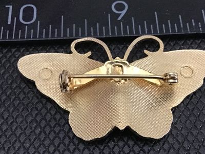 Sale Photo Thumbnail #183: A gold-tone butterfly brooch featuring purple translucent enamel wings accented with colorful pink, light blue, and white specks, with a blue body. Measures approximately 1.5 inches wide. Features a standard pin back with safety clasp in good working orde