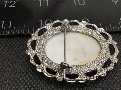 Sale Photo Thumbnail #173: An oval vintage brooch featuring a printed image of a London police officer (Bobby) in front of a red double-decker bus. Set in a silver-tone filigree metal frame with a pin-back closure. Shows light wear consistent with age.