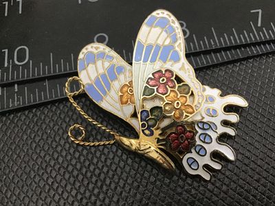 Sale Photo Thumbnail #54: A vintage gold-tone cloisonné enamel butterfly brooch pin featuring floral motifs on the wings in shades of blue, white, gold, green, and red. The back features a textured gold-tone finish with a standard pin stem and rollover safety clasp in working con