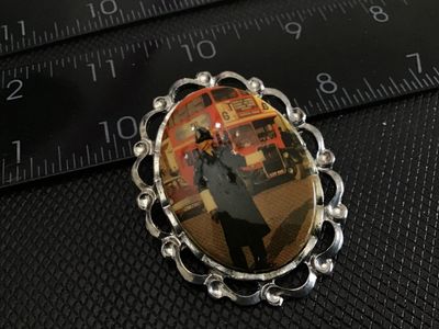 Sale Photo Thumbnail #172: An oval vintage brooch featuring a printed image of a London police officer (Bobby) in front of a red double-decker bus. Set in a silver-tone filigree metal frame with a pin-back closure. Shows light wear consistent with age.