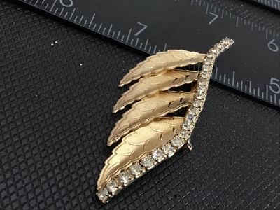 Sale Photo Thumbnail #146: A vintage gold-tone brooch featuring a cascading layered leaf motif with textured detail, accenting a curved line of clear set rhinestones along the top edge. The piece measures approximately 2.5 inches in length. Features a classic pin-back closure with 