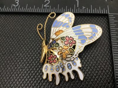 Sale Photo Thumbnail #56: A vintage gold-tone cloisonné enamel butterfly brooch pin featuring floral motifs on the wings in shades of blue, white, gold, green, and red. The back features a textured gold-tone finish with a standard pin stem and rollover safety clasp in working con