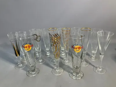 Sale Photo Thumbnail #54: Collection of assorted glassware and champagne flutes, featuring two Hard Rock Cafe Chicago pilsner-style glasses, gold-accented musical note and ribbon stems, and etched floral crystal flutes.