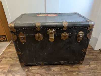 Sale Photo Thumbnail #471: Antique black flat-top steamer trunk featuring brass latches, hardware, and decorative corner brackets.