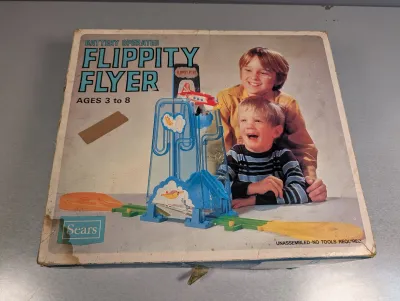 Sale Photo Thumbnail #211: Vintage Sears battery-operated Flippity Flyer toy set in its original box. The box shows age-related shelf wear, creases, and tape residue.