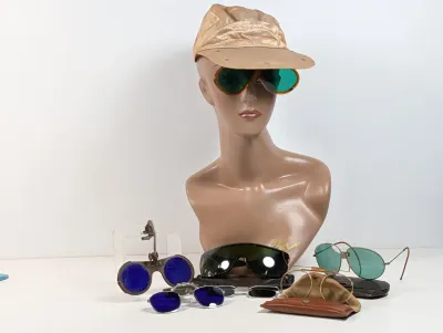 Sale Photo Thumbnail #103: Collection of vintage eyewear including clip-on sunglasses, aviators, safety goggles, and wire-rim glasses, displayed alongside a mannequin bust wearing a cap.
