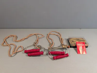 Sale Photo Thumbnail #385: Vintage exercise equipment set including two chest expanders with red wooden handles and bungee cords, accompanied by an Executive Exerciser jump rope in its original plastic case with instruction booklet.