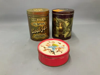 Sale Photo Thumbnail #354: Group of three decorative vintage tin canisters, including a Homestead Cookies tin, a 1992 Barcelona Olympic Games limited edition tin, and a round red tin featuring children and a dog.