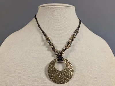 Sale Photo Thumbnail #2: Artisanal-style necklace featuring a large, hammered brass-tone circular pendant suspended on multi-strand dark cords with metallic bead accents.