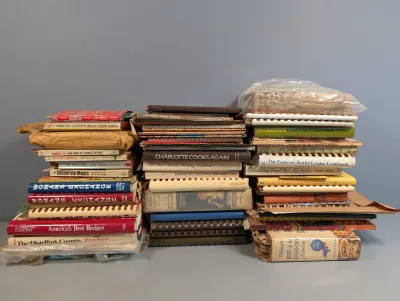 Sale Photo Thumbnail #287: Large collection of vintage mid-century cookbooks and recipe booklets stacked in three piles, including spiral-bound, hardcover, and paperback editions.