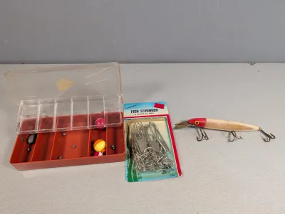 Sale Photo Thumbnail #317: Assorted fishing gear including a vintage wooden lure, a Sportfisher 9-hook fish stringer in original packaging, and an organizer box with weights and bobbers.