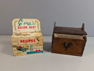 Sale Photo Thumbnail #250: Pair of vintage recipe boxes, including a mid-century printed metal tin reading 'Cora's Recipe Rest' and a dark wooden box featuring a metal rooster applique.