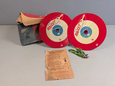 Sale Photo Thumbnail #275: Vintage Magne-Rocket Launcher game set by Toy Enterprises of America, complete with two target discs, feathered darts, original box, and instruction paper.