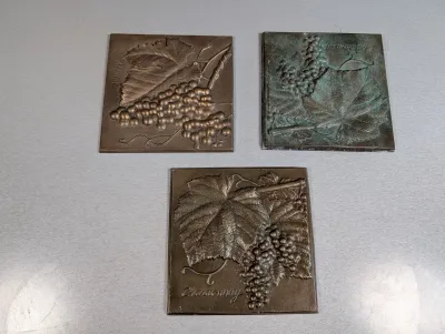 Sale Photo Thumbnail #48: Set of three square bronze bas-relief decorative plaques featuring grape clusters and vine leaves labeled Merlot and Chardonnay, showing varied patinas.