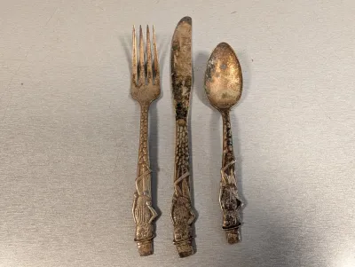 Sale Photo Thumbnail #252: Three-piece vintage brass or bronze flatware set featuring stylized tribal or African figural handles, showing heavy patina and age-related tarnish.