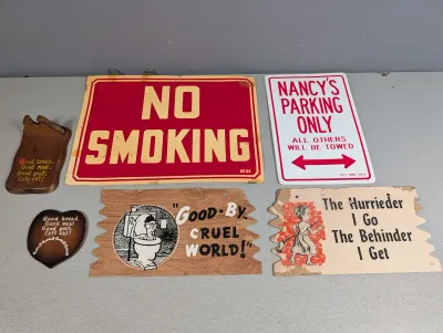 Sale Photo Thumbnail #319: Collection of vintage novelty and warning signs, including a red cardboard "No Smoking" sign, a printed metal "Nancy's Parking Only" sign, and various wooden wall plaques.