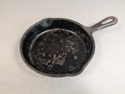 Sale Photo Thumbnail #302: Small vintage cast iron skillet with dual pour spouts, showing light surface rust and residue that requires cleaning and re-seasoning.